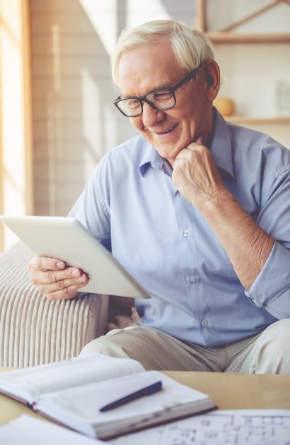 Handsome old man in eyeglasses is using a digital tablet and smiling while sitting on couch at home